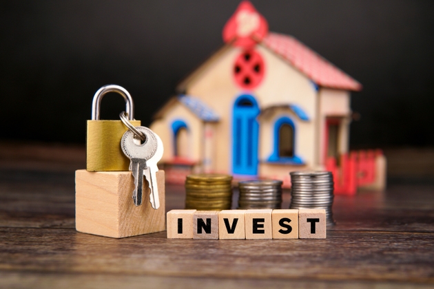 Padlock and keys placed on wooden block beside stacked coins and wooden cubes spelling INVEST, with a miniature house in the background. Concept of real estate investment, property ownership, and financial security.