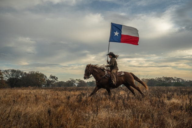 Woman Wearing Cowboy Hat Riding Running Horse While Holding Large Texas Flag In Grassy Field With Cloudy Sky