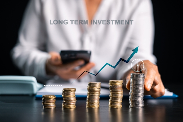 Long term investment concept with rising coin stacks and upward chart as person uses smartphone at desk, symbolizing compound growth, savings plan, time horizon and financial progress. savings plan
