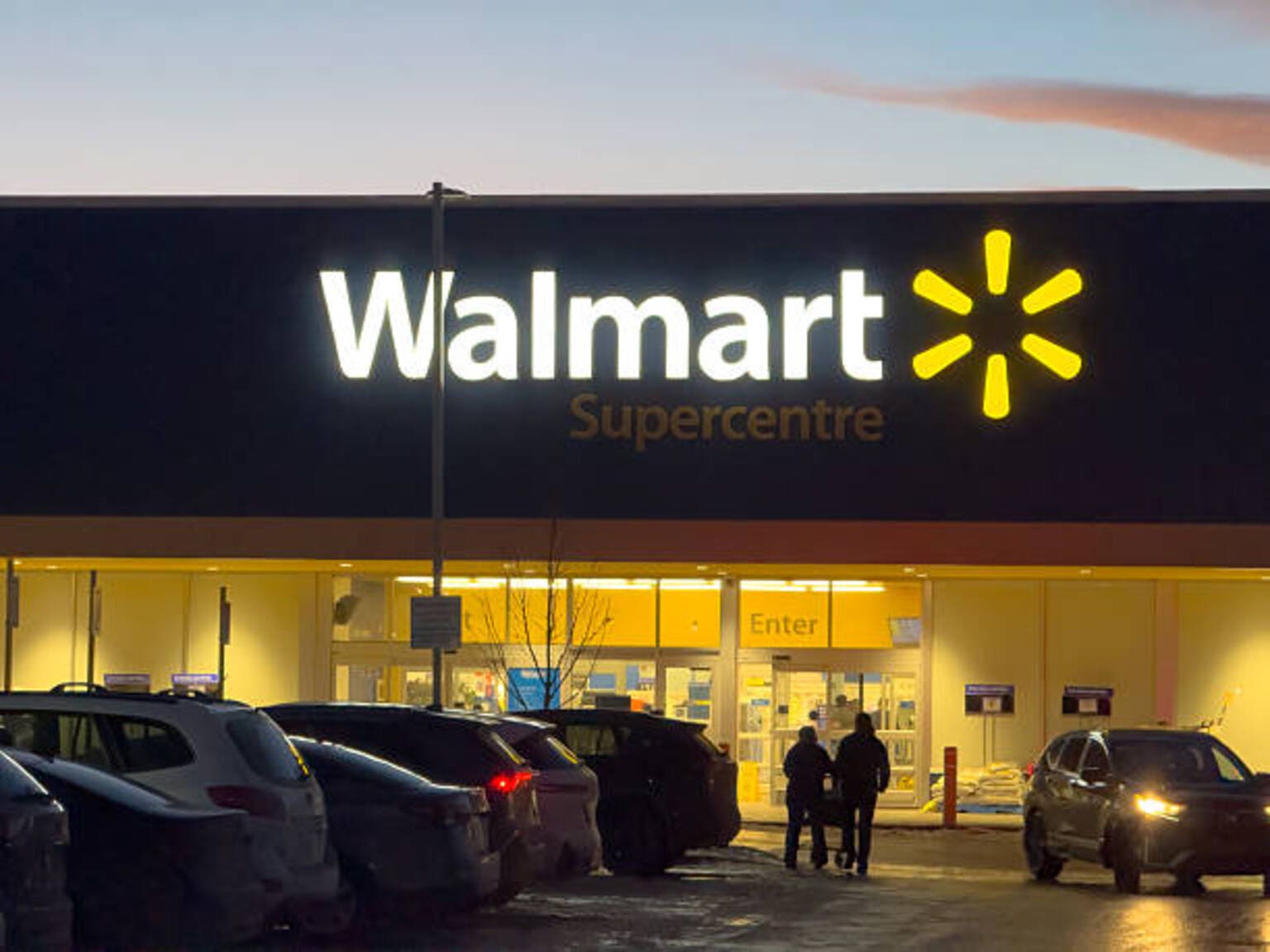 A Walmart Supercentre storefront with an illuminated brand logo and customers entering the building at dusk.