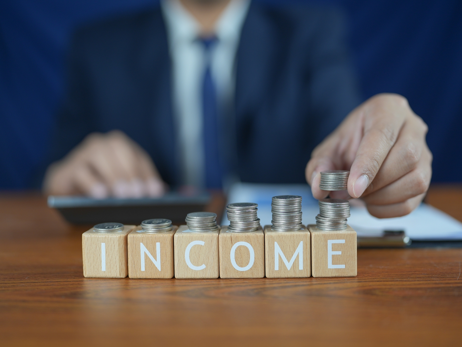Person Counting Coins and Stacking Them on Wooden Blocks Spelling Income with Calculator and Clipboard in Background