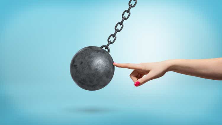 A giant female arm with one finger lightly touching a black iron wrecking ball and stopping its movement.