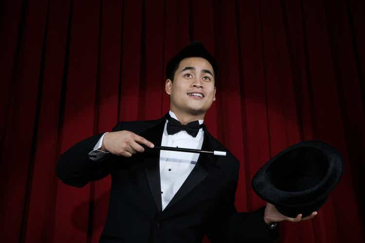 Asian magician in tuxedo pointing to top hat with wand