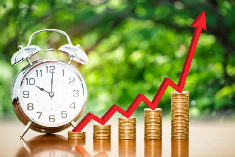 Stack of money coins arranged as a graph with red arrow indicates economic growth and alarm clock on a wooden table with blurred background, time to money growth concept