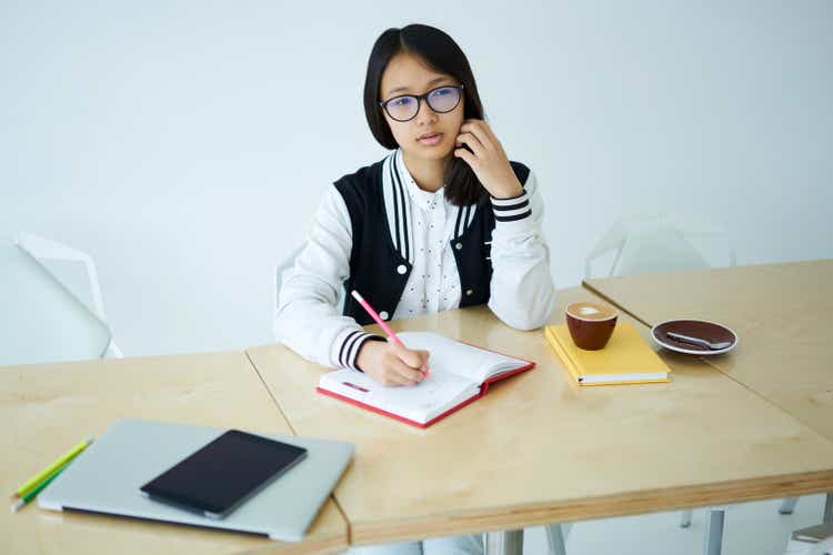 Thoughtful chinese school teenager thinking about idea to write in notebook while preparing for college exams.Asian hipster girl dressed in trendy wear doing notes for creating article of new web blog