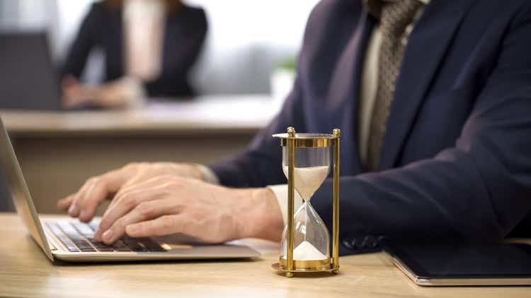 Businessman typing on laptop at desk, hourglass trickling, deadline approaching