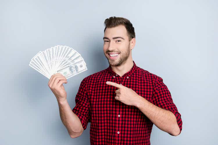 Portrait of handsome confident with toothy beaming smile smart rich wealthy friendly cheerful excited entrepreneur demonstrating many banknotes in hand with forefinger isolated on gray background