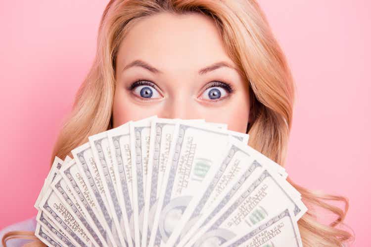 Close up portrait of shocked surprised girl peeking out fan of much money with wide open eyes hiding her half face isolated on pink background