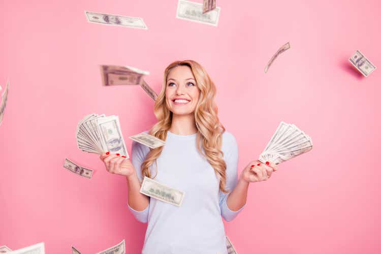 Portrait of pretty charming positive cute successful lucky cheerful girl standing under shower from money having a lot of money in hands isolated on pink background