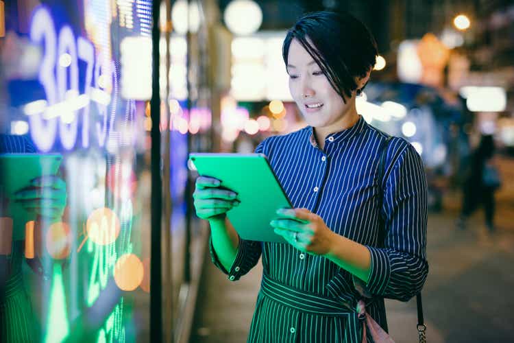 Asian businesswomen checking stock market data on tablet before Hong Kong financial display board