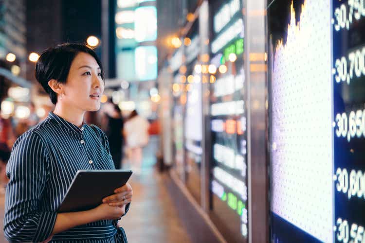 Asian businesswomen checking stock market data on tablet before Hong Kong financial display board