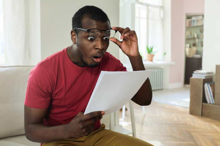 Young african american man sitting on couch in modern apartment with mouth open, holding utility bill with high rates, raised eyeglasses in wow or surprise gesture