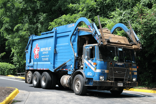A blue garbage truck with a crane in the back Description automatically generated