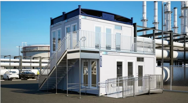 A two-story modular office building (WillScot) situated on an industrial site, featuring a staircase and ramp access, with large storage tanks and piping visible in the background.