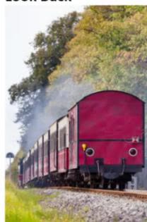 A photograph of a red freight train moving from left to right, with a large plume of white smoke trailing behind it. The train is on a track with green grass and trees in the background.