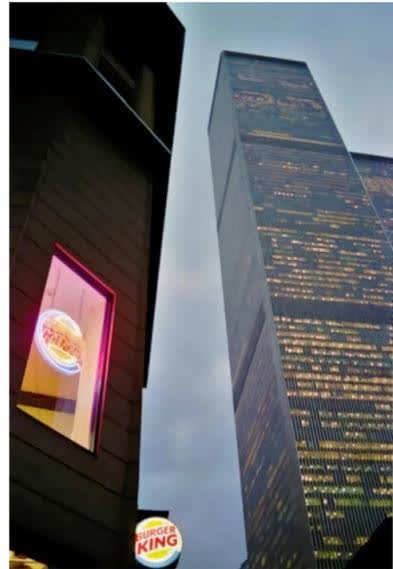 A photograph of a Burger King restaurant building at night, with its illuminated sign visible. In the background, the tall, modern skyscraper of the World Trade Center is visible, also illuminated. The scene is set in an urban environment with other buildings and streetlights in the distance.