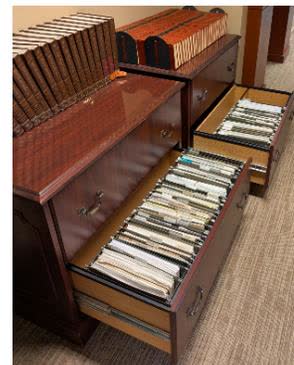 A photograph of a wooden filing cabinet with several drawers open, revealing stacks of papers and folders, illustrating the 'analog' research records mentioned in the text.