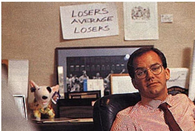 A photograph of Paul Tudor Jones sitting at his desk. Behind him, a sign on the wall reads 'LOSERS AVERAGE LOSERS'. To his left, a small stuffed animal is visible on the desk.